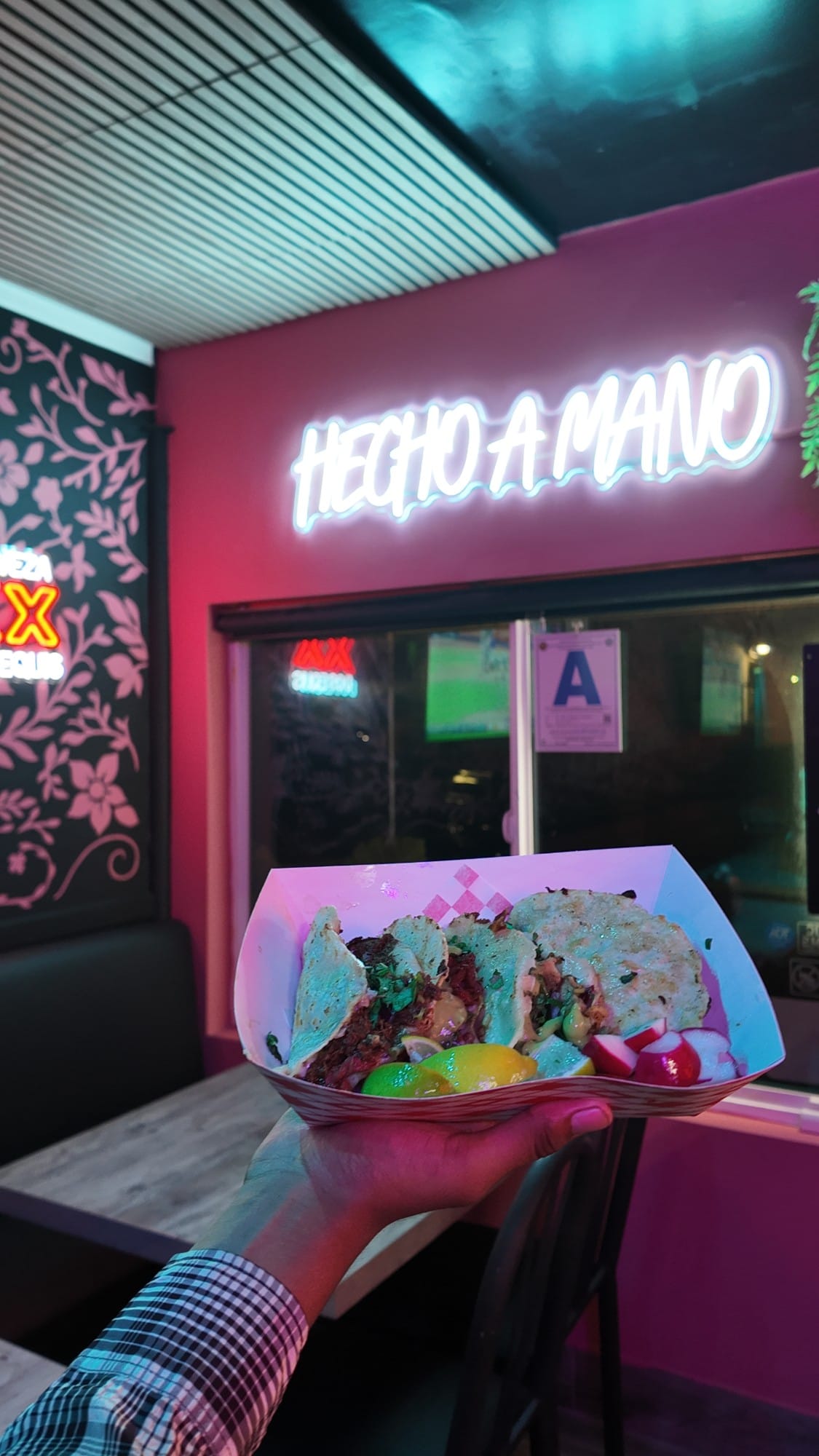 A hand holding a tray of tacos with lime wedges and sliced radishes in front of the glowing "Hecho a mano" neon sign inside Los 3 Animales