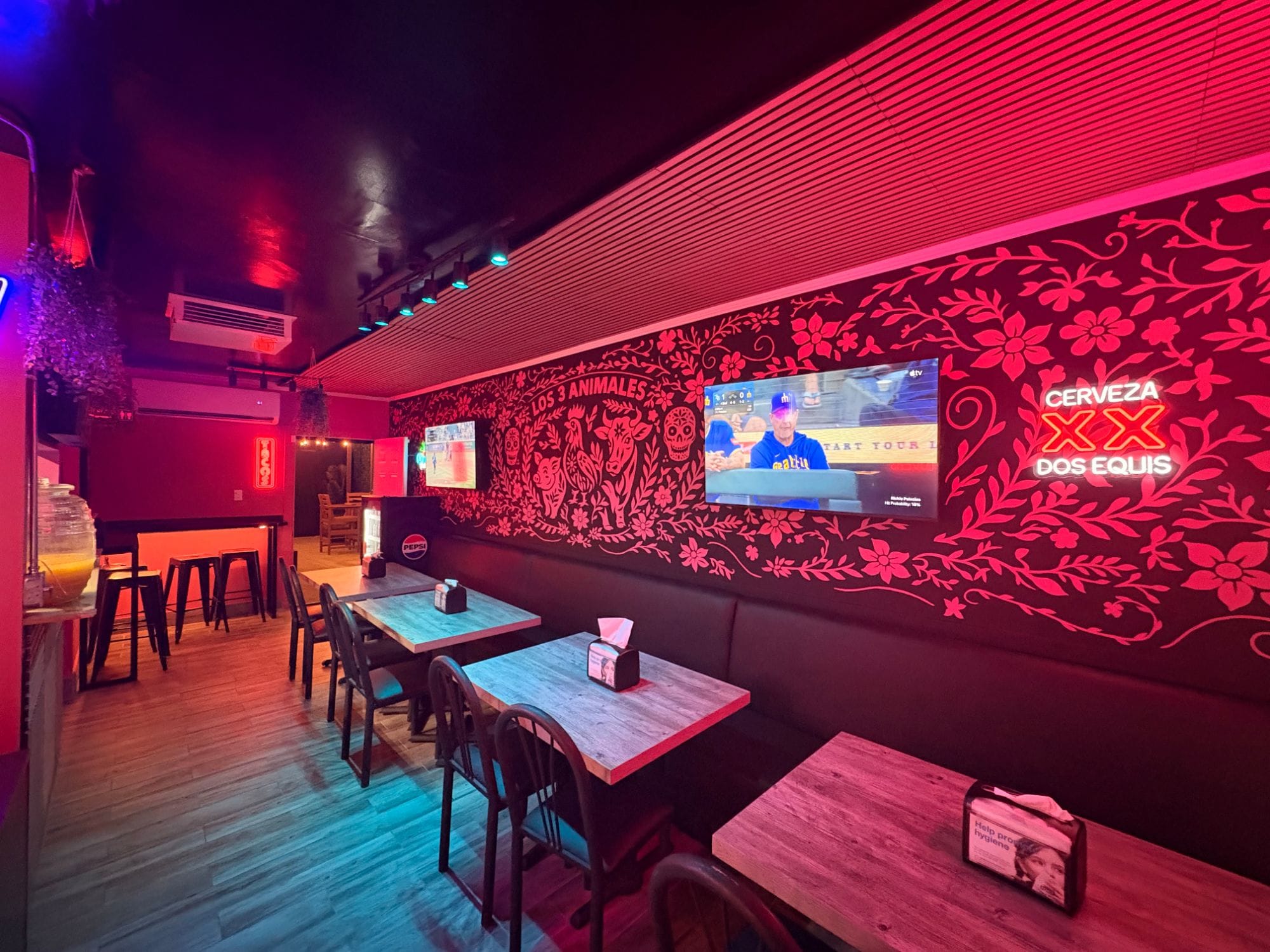The Los 3 Animales dining room glowing pink: the hand-painted pig, rooster, and bull mural on a black wall, a long booth with wooden tables, TVs, and neon signs
